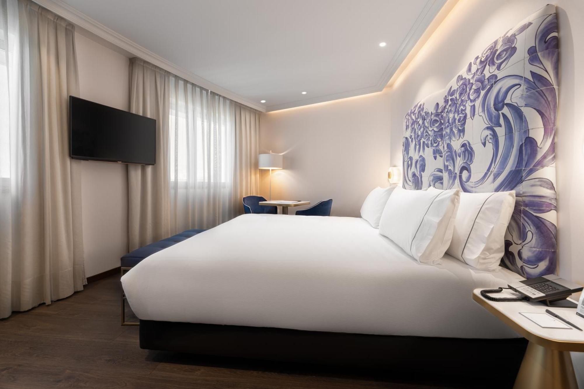 Eurostars Universal Lisboa Dış mekan fotoğraf The photo shows a modern hotel room featuring a large bed with white bedding and several decorative pillows. Behind the bed, there is a decorative wall with a blue floral design. To the left, there is a flat-screen TV mounted on the wall, and near th