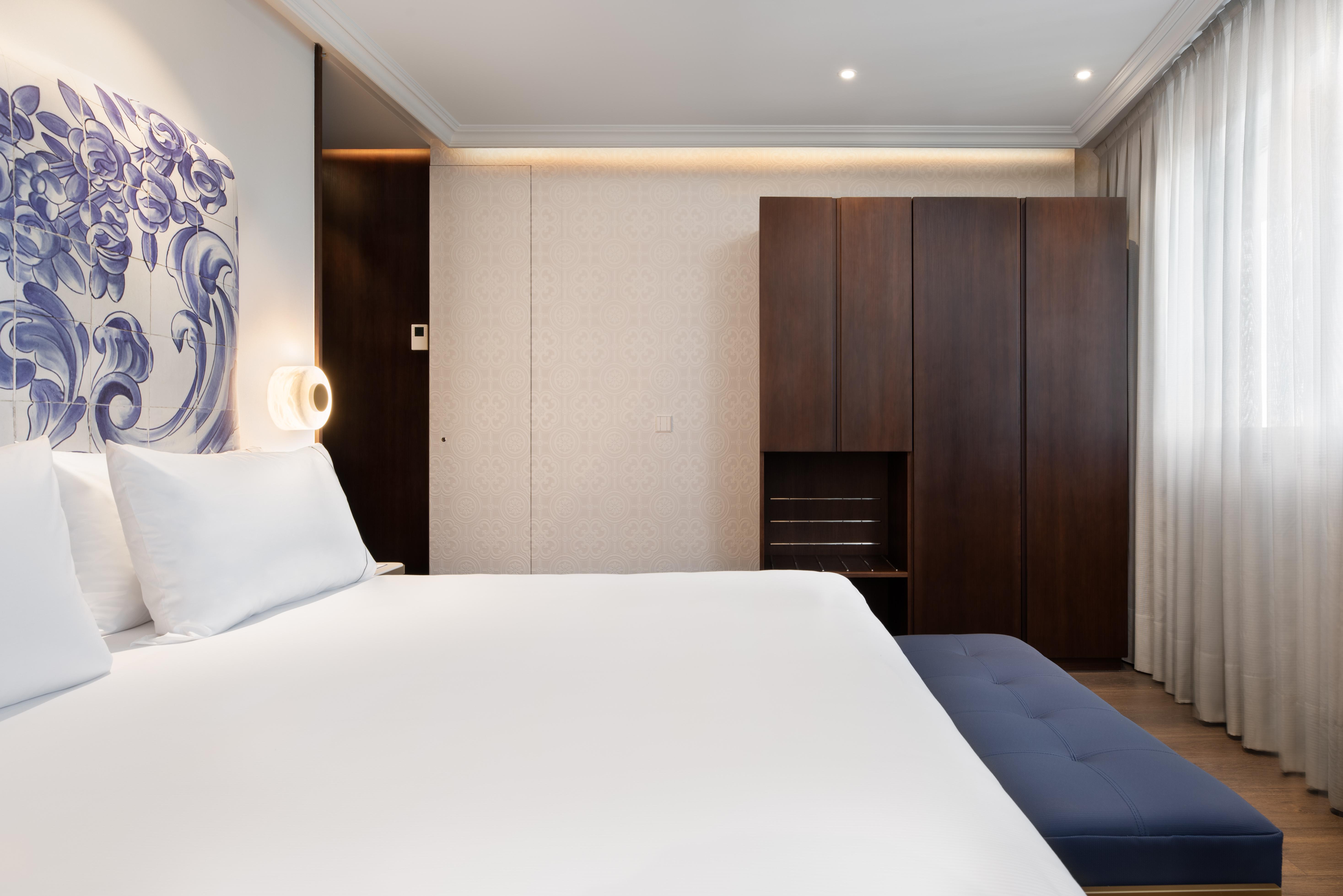 Eurostars Universal Lisboa Dış mekan fotoğraf The photo depicts a modern hotel room with a contemporary design. It features a large bed with crisp white linens and two pillows. Behind the bed, there is a decorative blue and white wall art. To the side, there is a dark wood wardrobe or cabinet, a