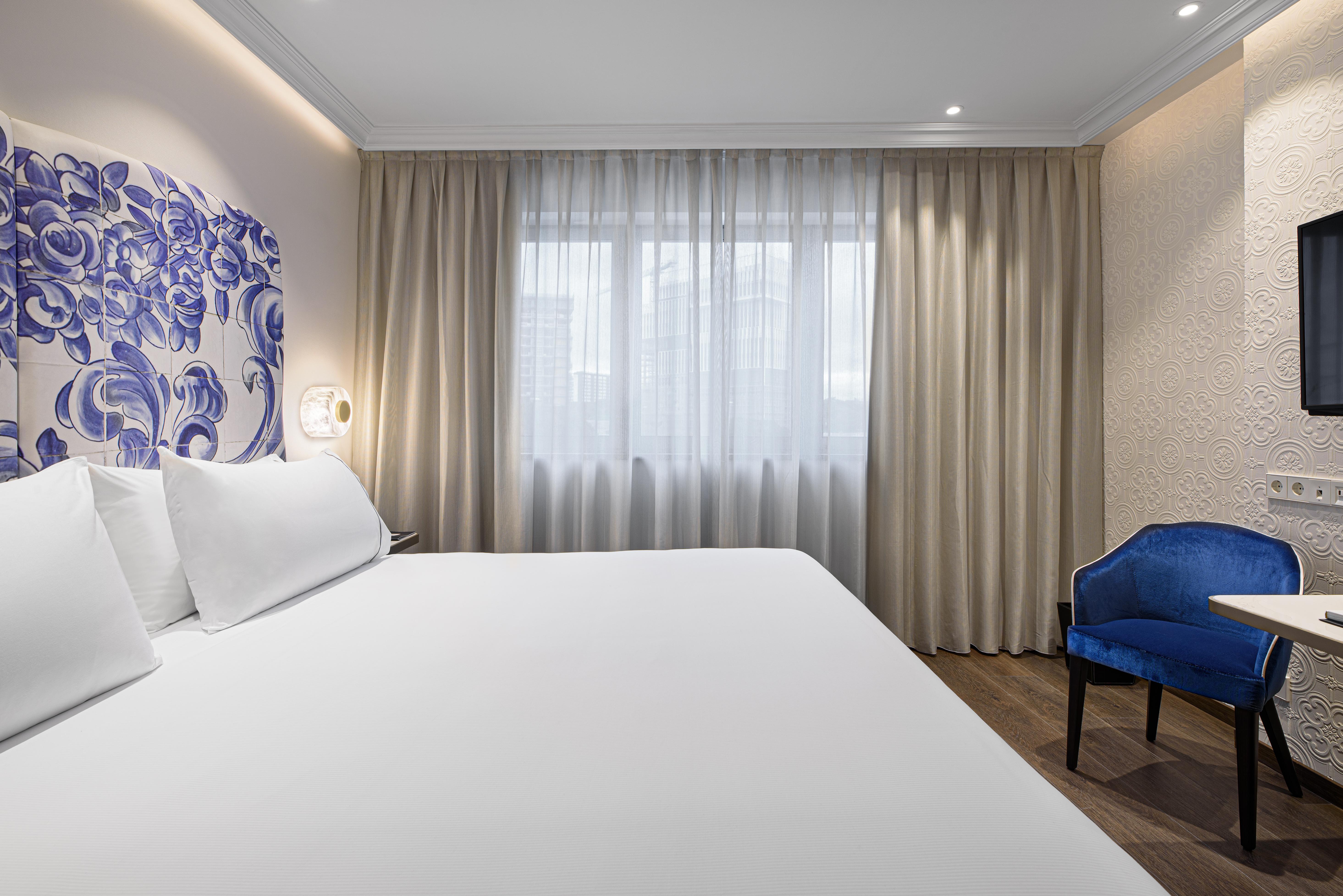 Eurostars Universal Lisboa Dış mekan fotoğraf The photo shows a modern hotel room with a minimalistic design. It features a large bed with white linens and two pillows. Behind the bed, there is a decorative wall with a blue floral design. To the side, a window is covered with sheer curtains, all