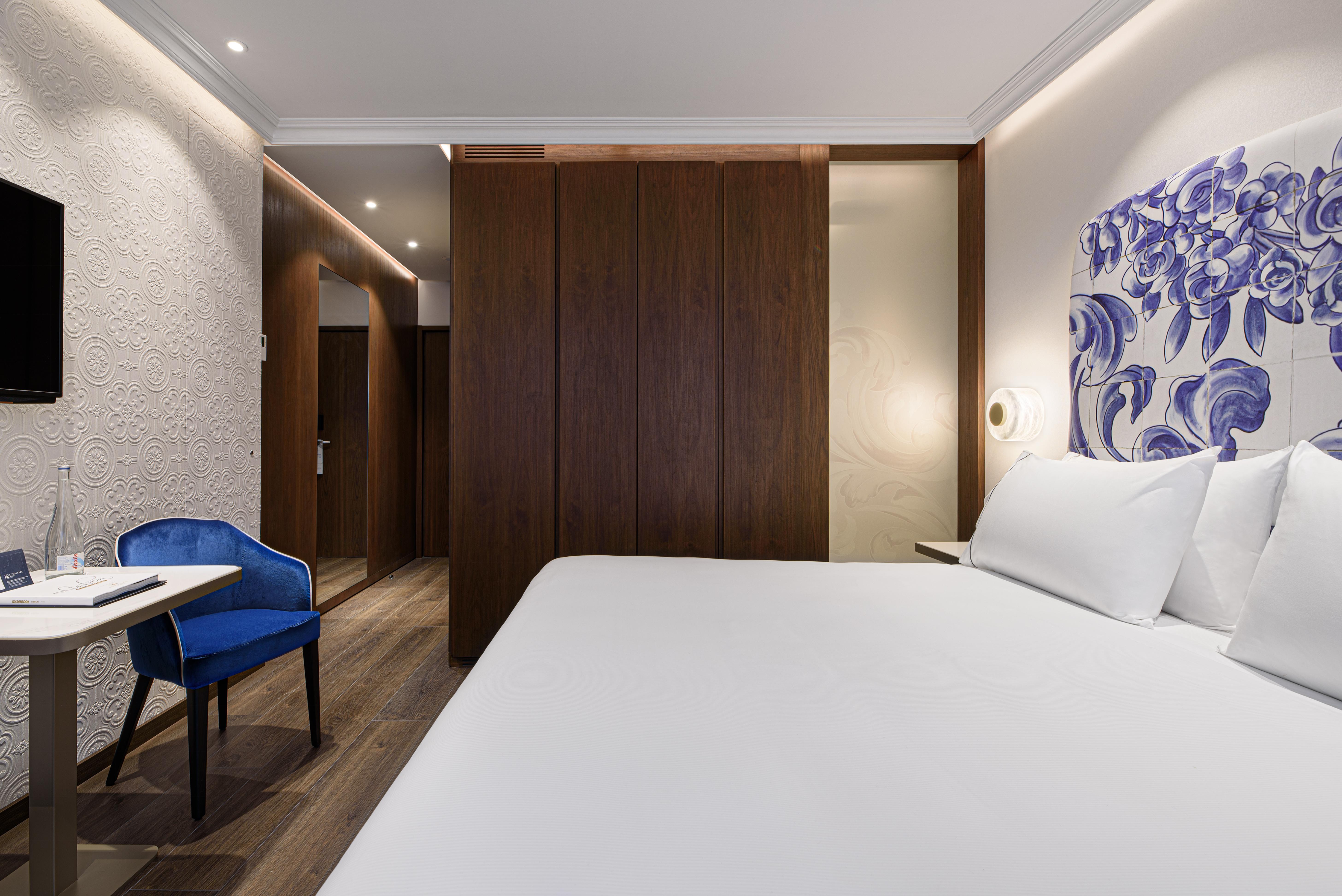 Eurostars Universal Lisboa Dış mekan fotoğraf The photo shows a modern hotel room. It features a large bed with white linens, positioned against a wall with decorative artwork. To the left, there is a small table and a blue chair, adding a pop of color to the neutral tones of the room. The light