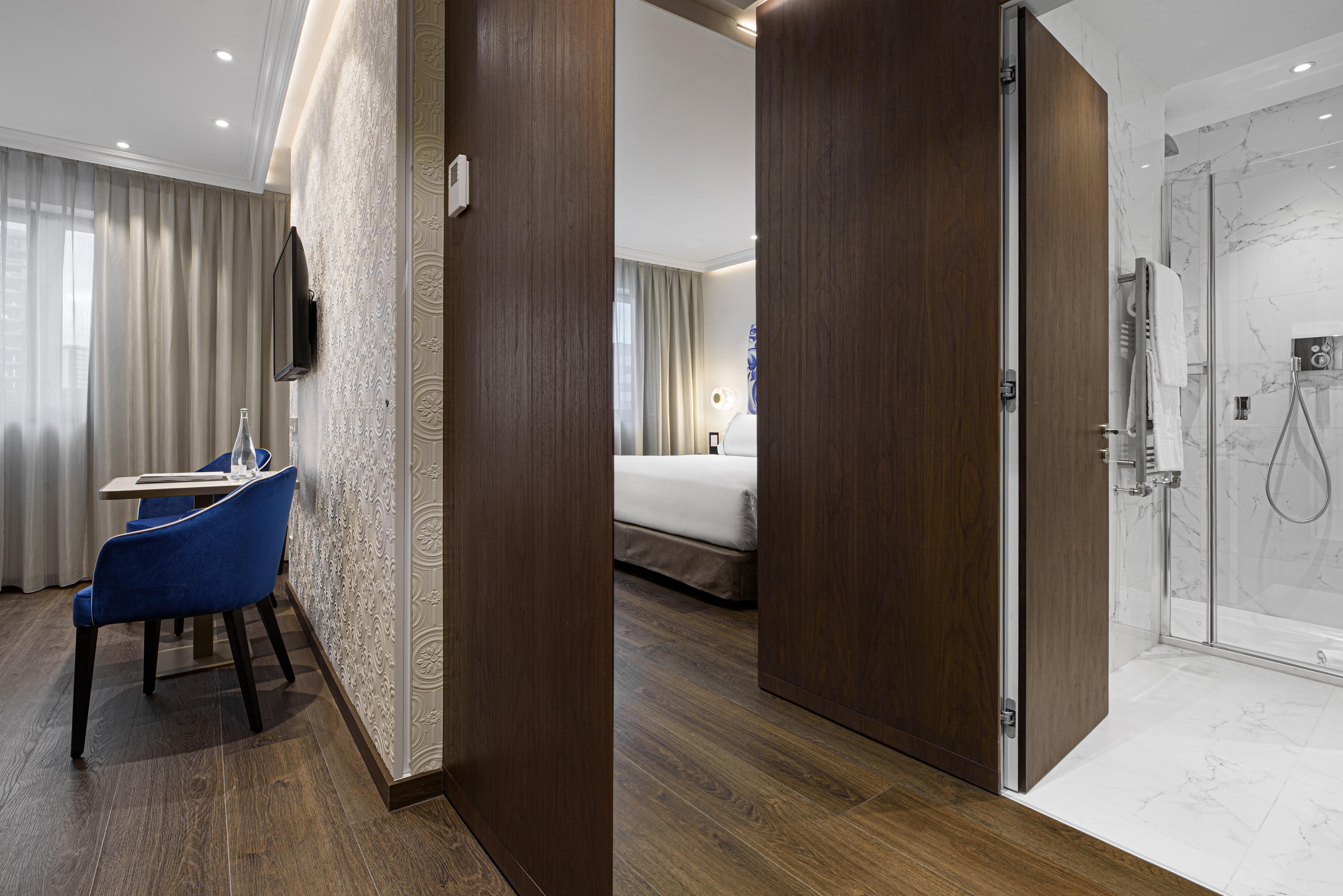 Eurostars Universal Lisboa Dış mekan fotoğraf The photo shows the interior of a hotel room. In the foreground, there are two partially open wooden doors leading into the room. To the left, a small desk with a blue chair is visible, along with a wall-mounted television. The room features light-co