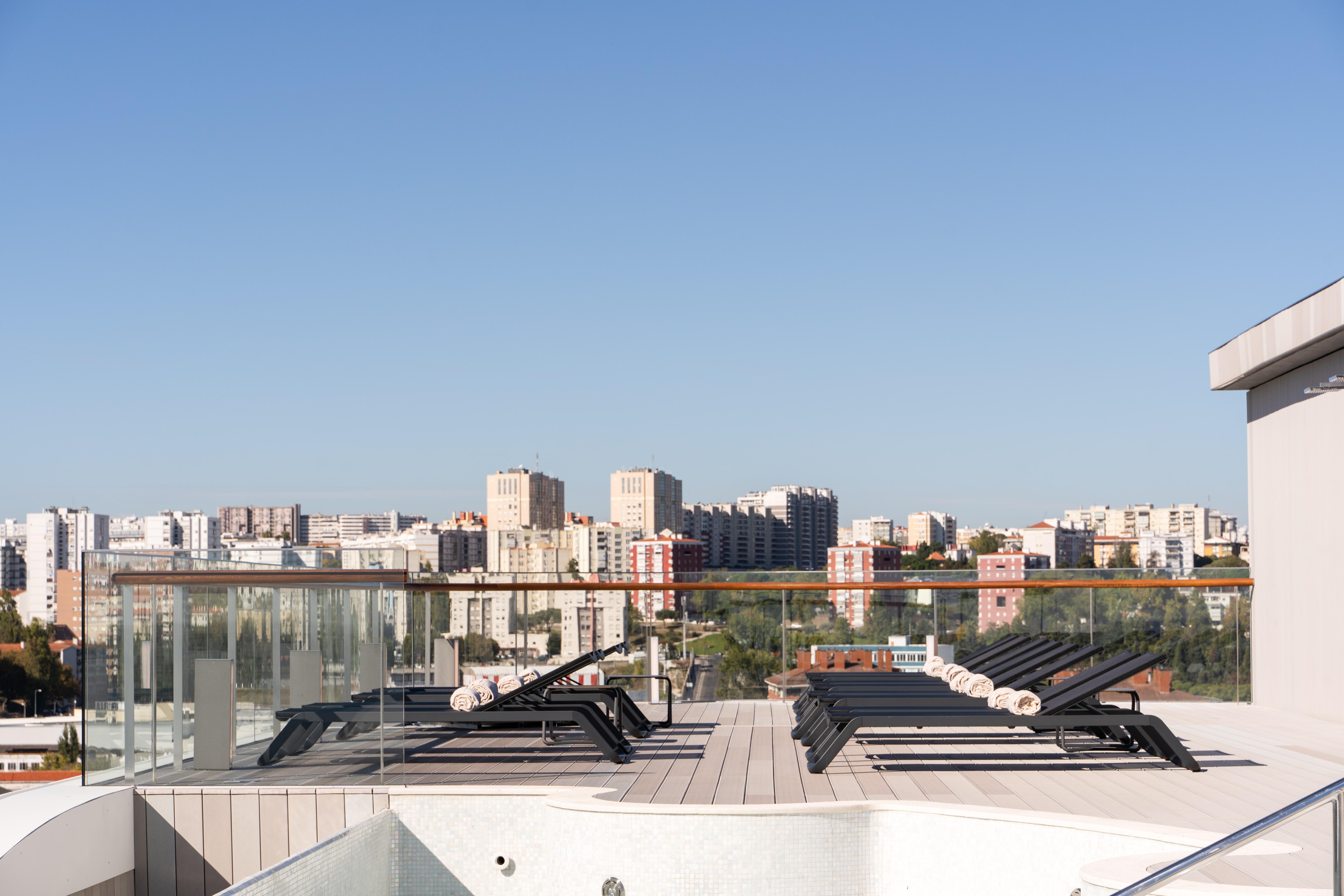 Eurostars Universal Lisboa Dış mekan fotoğraf The image shows a rooftop setting with a clear blue sky. In the foreground, there are several lounge chairs arranged on a wooden deck. Beyond the deck, a transparent railing allows for an unobstructed view of a cityscape, which includes various build