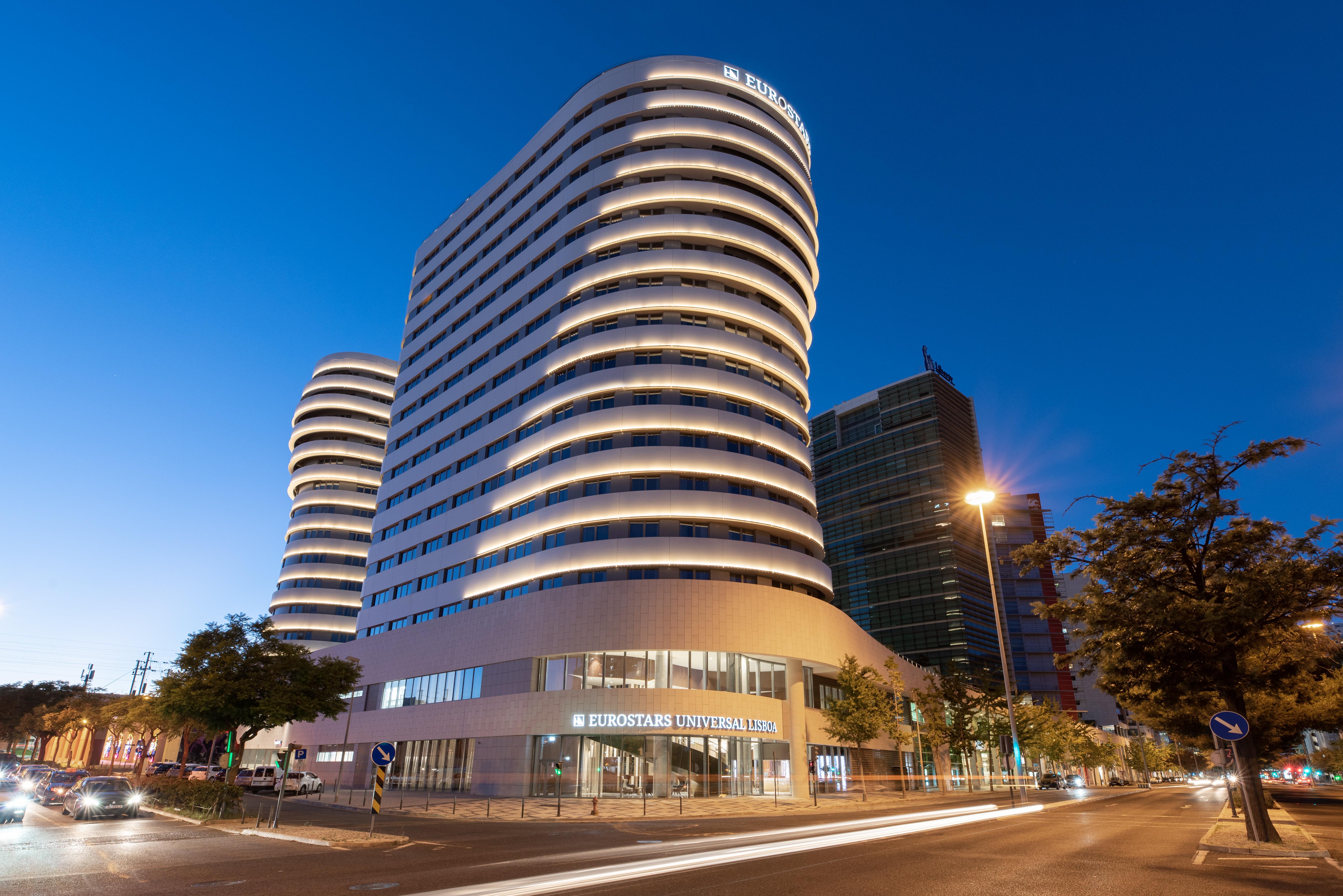 Eurostars Universal Lisboa Dış mekan fotoğraf The photo shows a modern building complex featuring two distinct towers. The towers have a smooth, curved facade with horizontal lines that create a sleek, contemporary look. The building is illuminated with soft lighting that highlights its architec