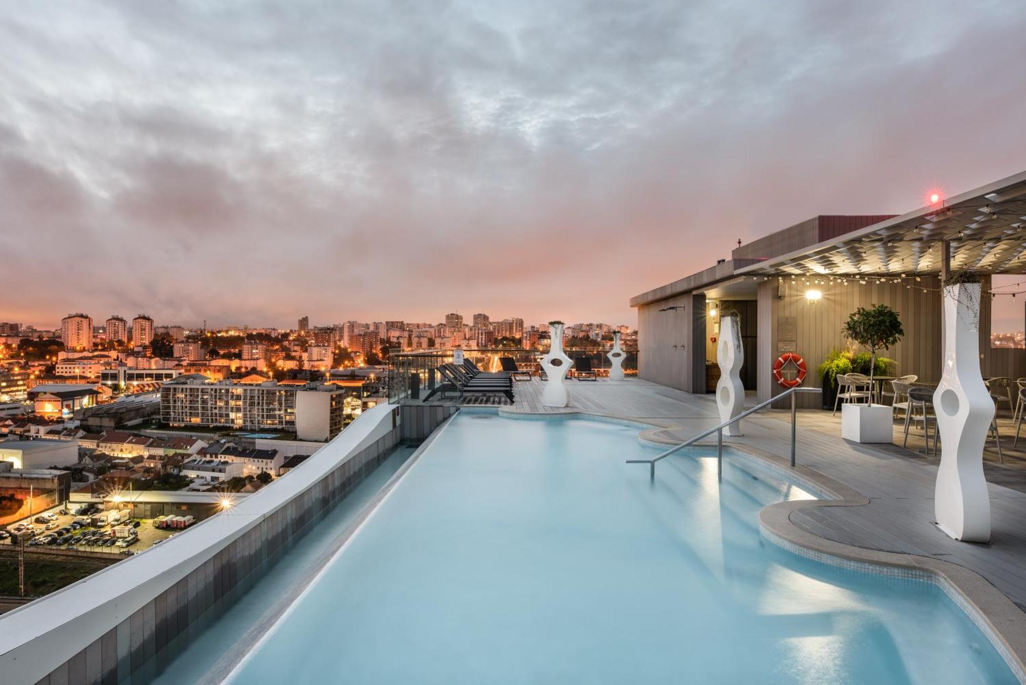 Eurostars Universal Lisboa Dış mekan fotoğraf The photo shows a rooftop pool with a modern design, featuring clear blue water and a sleek edge that appears to blend into the skyline. In the background, there are city buildings illuminated by sunset colors, giving the scene a warm and inviting at