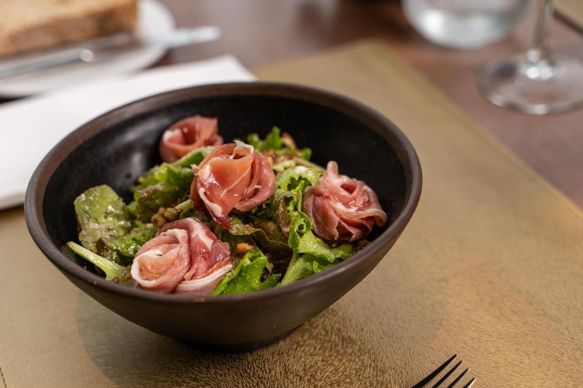 Eurostars Universal Lisboa Dış mekan fotoğraf The photo shows a bowl of salad with a mix of fresh greens, topped with rolled slices of cured meat, likely ham. The salad has a vibrant color, featuring leafy greens and the pinkish hue of the meat. The bowl is dark and round, with a simple, elegant