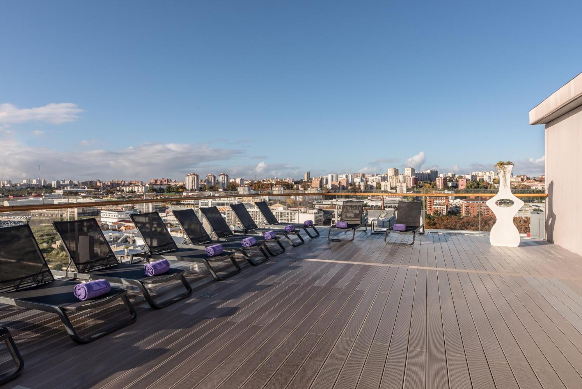 Eurostars Universal Lisboa Dış mekan fotoğraf The photo shows a rooftop terrace with several lounge chairs arranged in a row. Each chair is equipped with a purple towel. The terrace has a wooden deck and offers a panoramic view of a cityscape with various buildings in the background. The sky is 