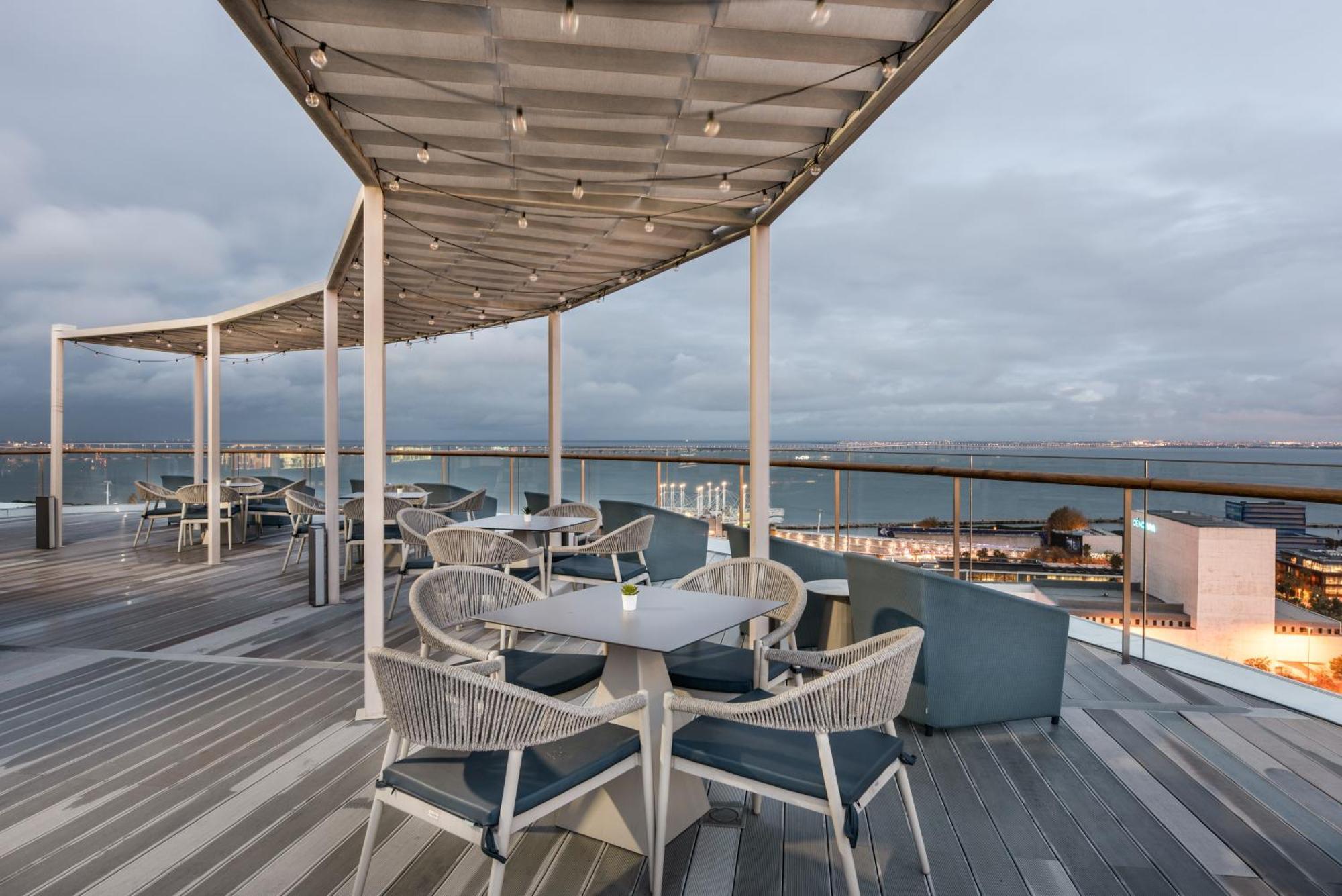 Eurostars Universal Lisboa Dış mekan fotoğraf The photo shows a scenic outdoor dining area with modern furniture. There are several round tables and chairs arranged on a wooden deck. Some of the tables have blue cushions, and the area is covered by a sleek, curved canopy that is adorned with str