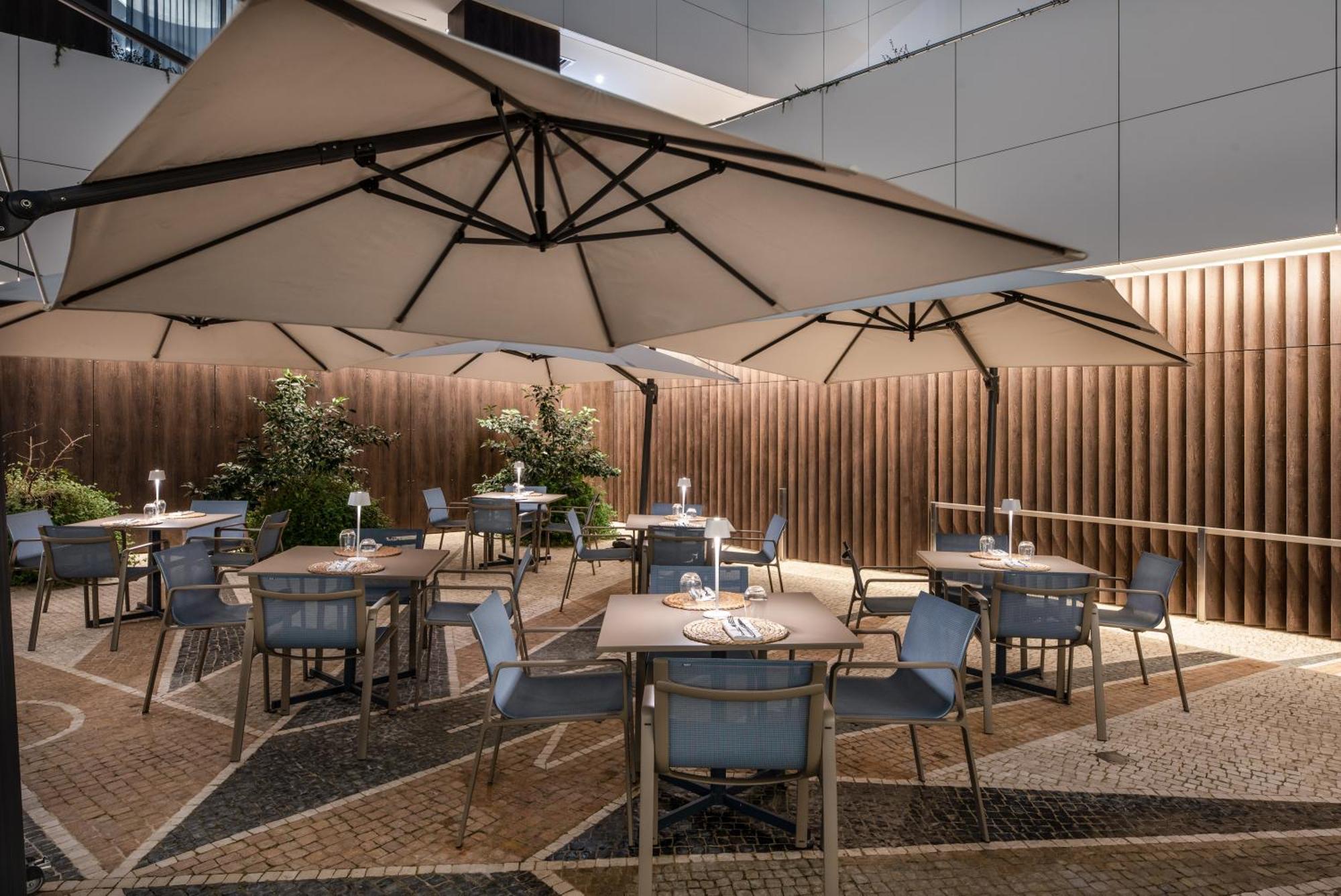 Eurostars Universal Lisboa Dış mekan fotoğraf The photo depicts an outdoor dining area featuring several tables and chairs arranged under large umbrellas. The setting is likely part of a restaurant or café, with a nicely designed stone or tiled floor. There are plants in the background, providin
