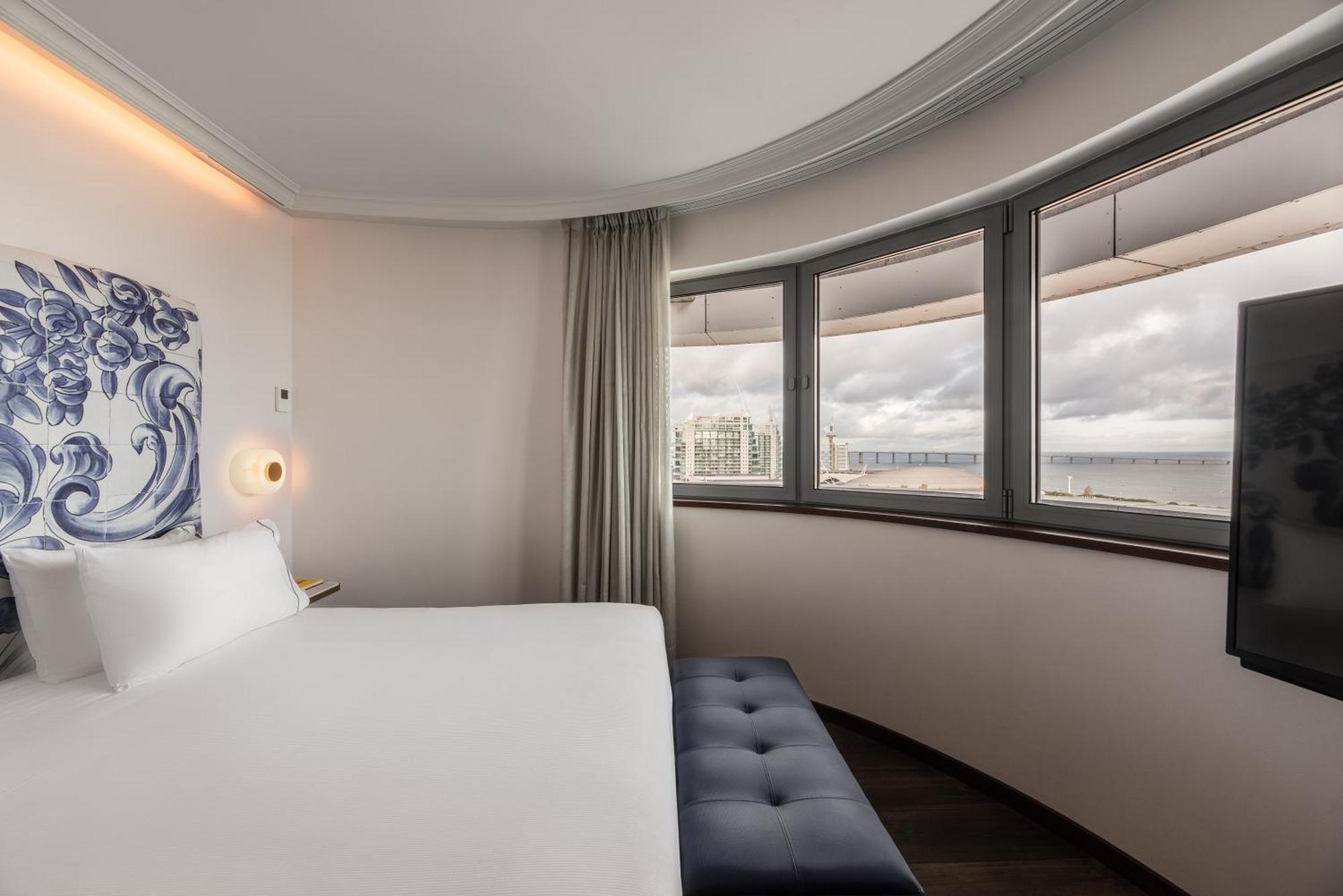 Eurostars Universal Lisboa Dış mekan fotoğraf The photo shows a modern hotel room with a curved wall design. It features a bed with white linens positioned against the wall. There is a stylish ottoman or bench at the foot of the bed. The room has large windows that provide a view, allowing natur