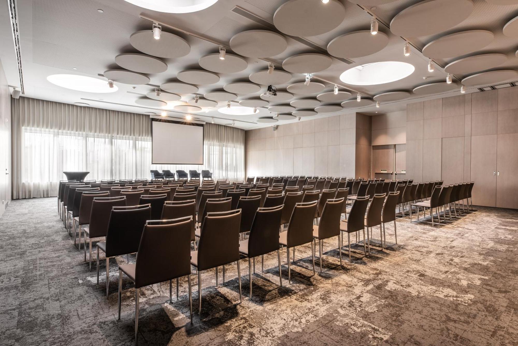 Eurostars Universal Lisboa Dış mekan fotoğraf The photo shows a conference or meeting room that is set up for an event. There are rows of brown chairs arranged facing a screen at the front of the room. The space has a modern design, with a decorative ceiling featuring circular elements. Large wi