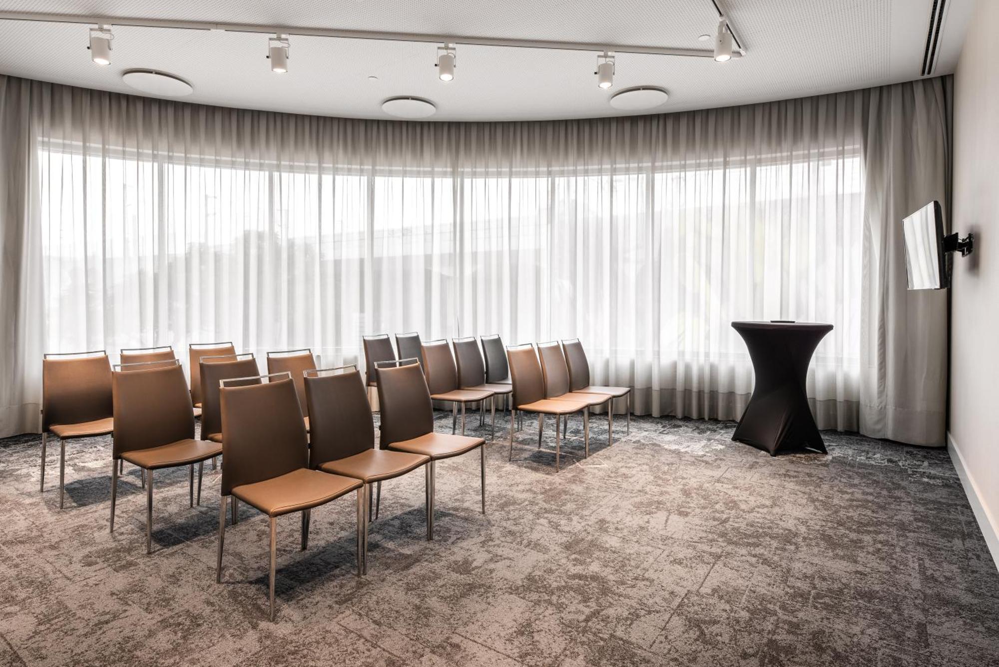 Eurostars Universal Lisboa Dış mekan fotoğraf The photo shows a modern meeting room with a curved design. There are several rows of simple, brown chairs arranged in the space, facing a small black podium. The room features large windows covered with sheer curtains, allowing natural light to filt