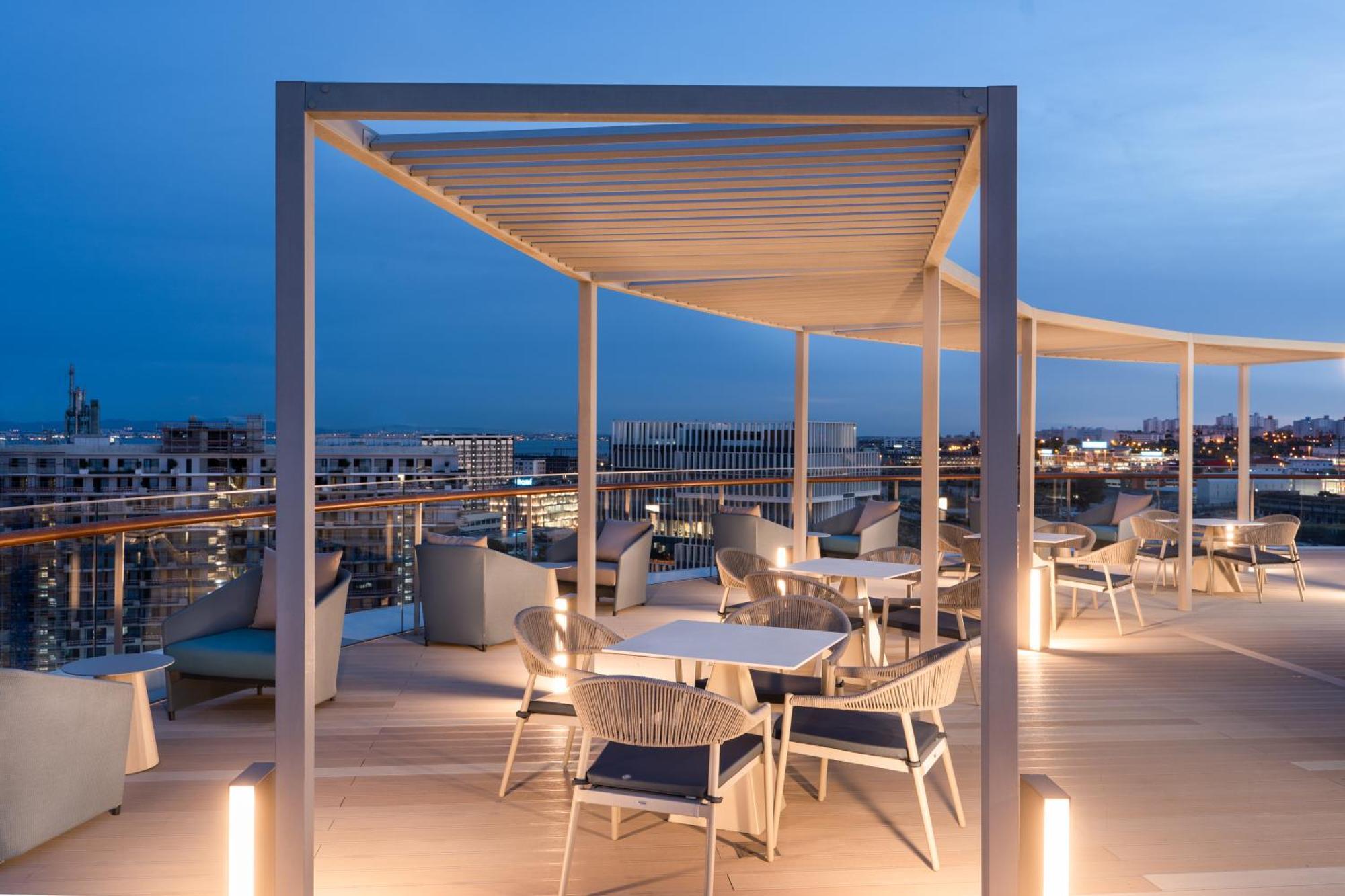 Eurostars Universal Lisboa Dış mekan fotoğraf The photo shows a stylish rooftop terrace area, likely part of a restaurant or lounge. It features modern outdoor furniture, including tables and chairs, arranged on a wooden deck. A white pergola provides shade overhead, and there are lights enhanci