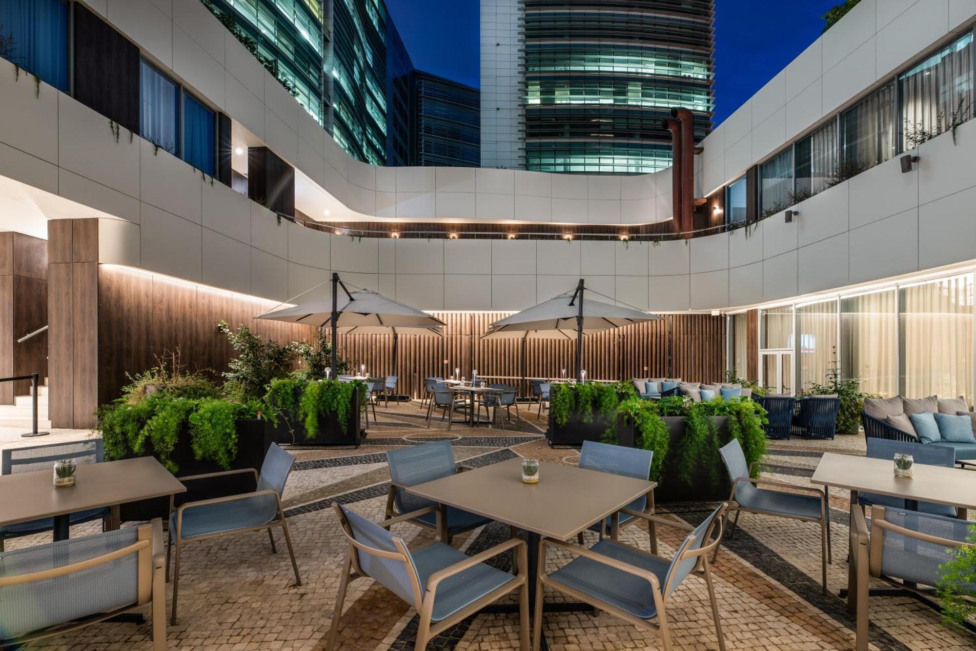 Eurostars Universal Lisboa Dış mekan fotoğraf The photo depicts an outdoor seating area likely within a modern urban setting, possibly a hotel or office complex. It features several tables and chairs arranged around the space, with large umbrellas providing shade. Lush greenery is seen in variou